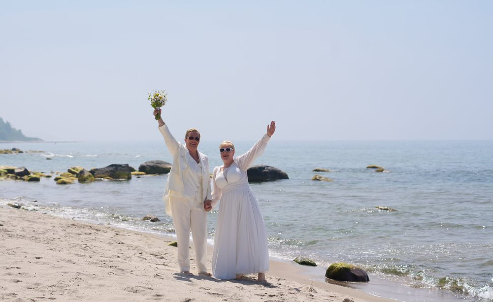 A just married lesbian couple standing on the beach in Bornholm and waving.