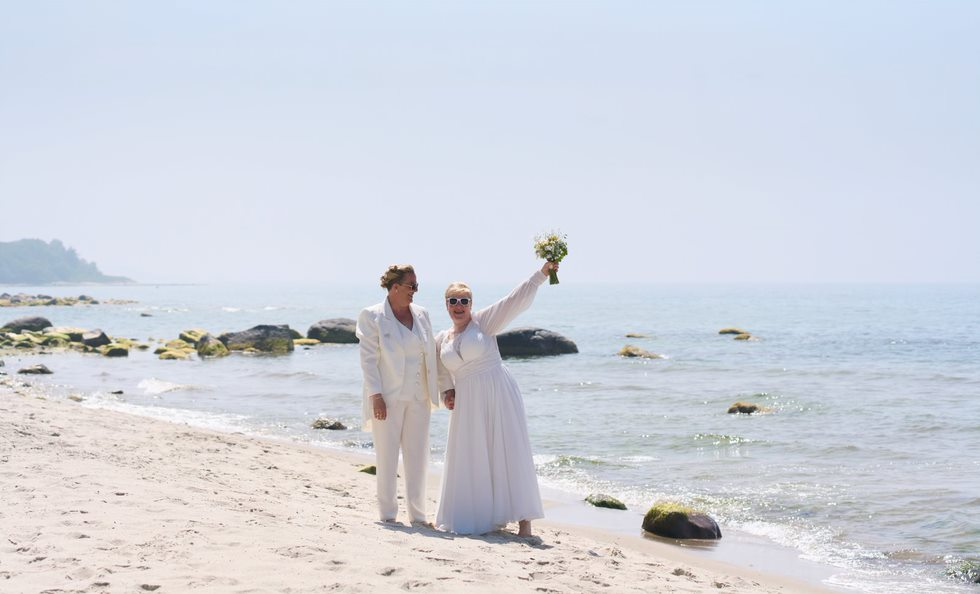 Same sex female couple waves at the wedding photographer during a beach photo shoot after their wedding on Bornholm in Denmark.